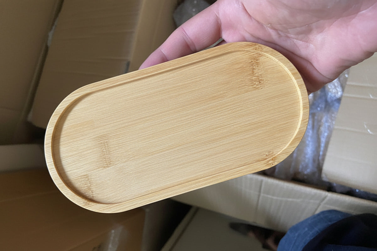 A hand holding a light natural bamboo oval tray with a raised rim, shown against a background of cardboard boxes and packaging materials.