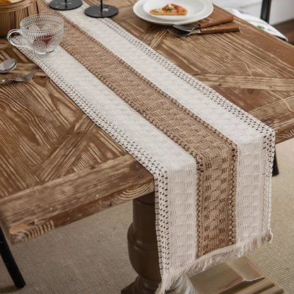 A woven table runner with a striped pattern in cream, beige, and brown, draped over a rustic wooden dining table. The runner features a textured knit design with fringe on one end.