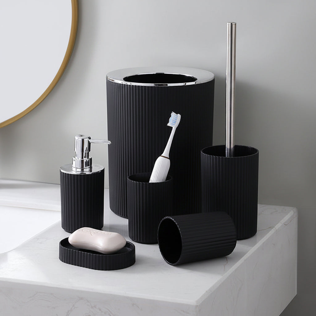 A bathroom accessory set in matte black ribbed plastic, including a toilet brush holder with a silver metal handle, a toothbrush holder, a soap dispenser, a bar of soap, and a small lid, arranged on a white marble countertop.
