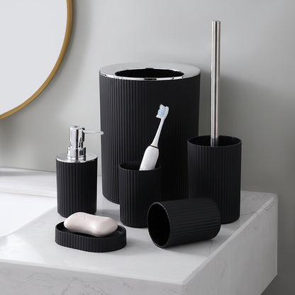A bathroom accessory set in matte black ribbed plastic, including a toilet brush holder with a silver metal handle, a toothbrush holder, a soap dispenser, a bar of soap, and a small lid, arranged on a white marble countertop.